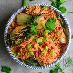 A ceramic bowl of noodles with vegetables and herbs.