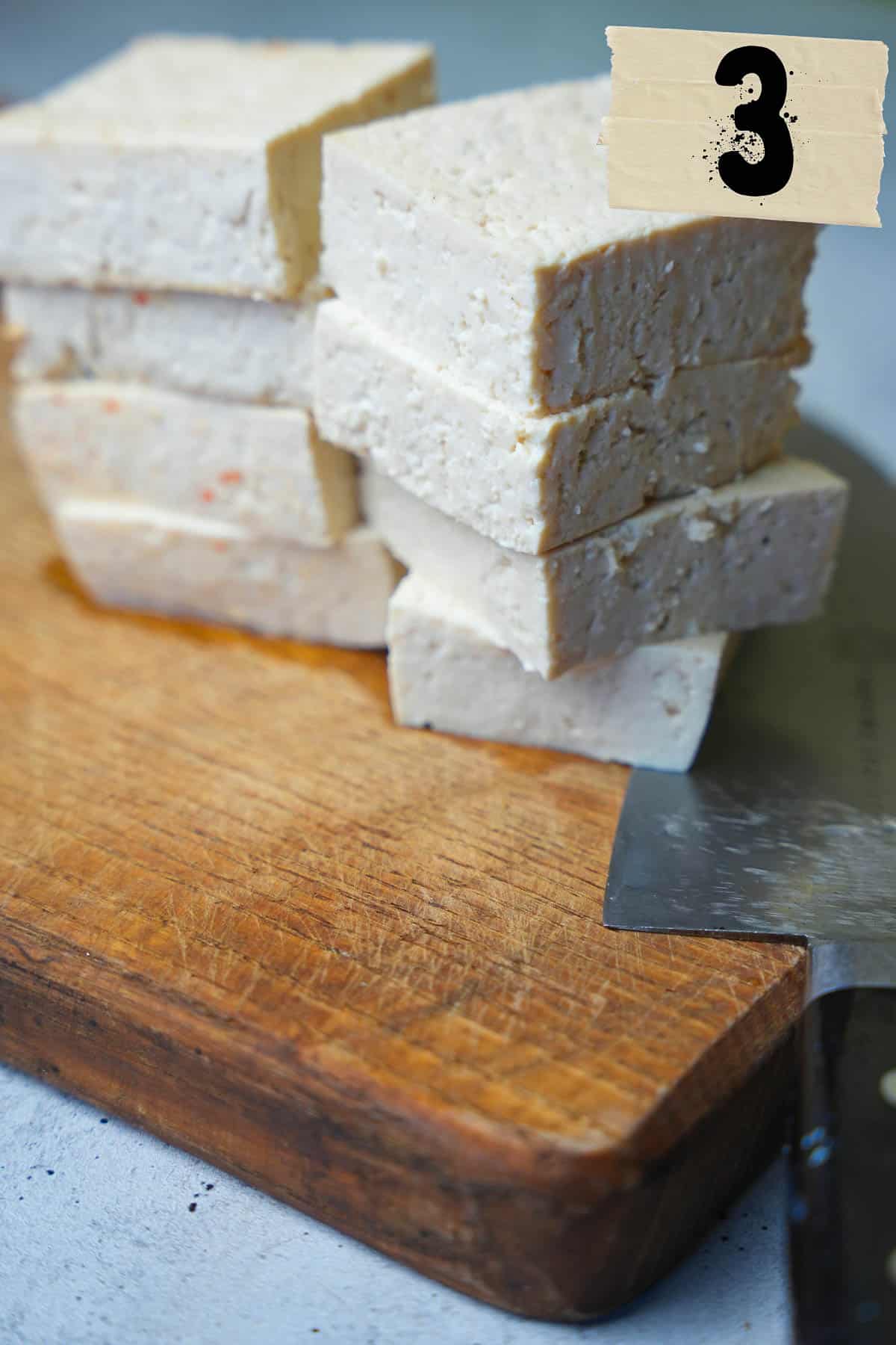 A block of tofu cut into eight pieces on a cutting board next to a knife.
