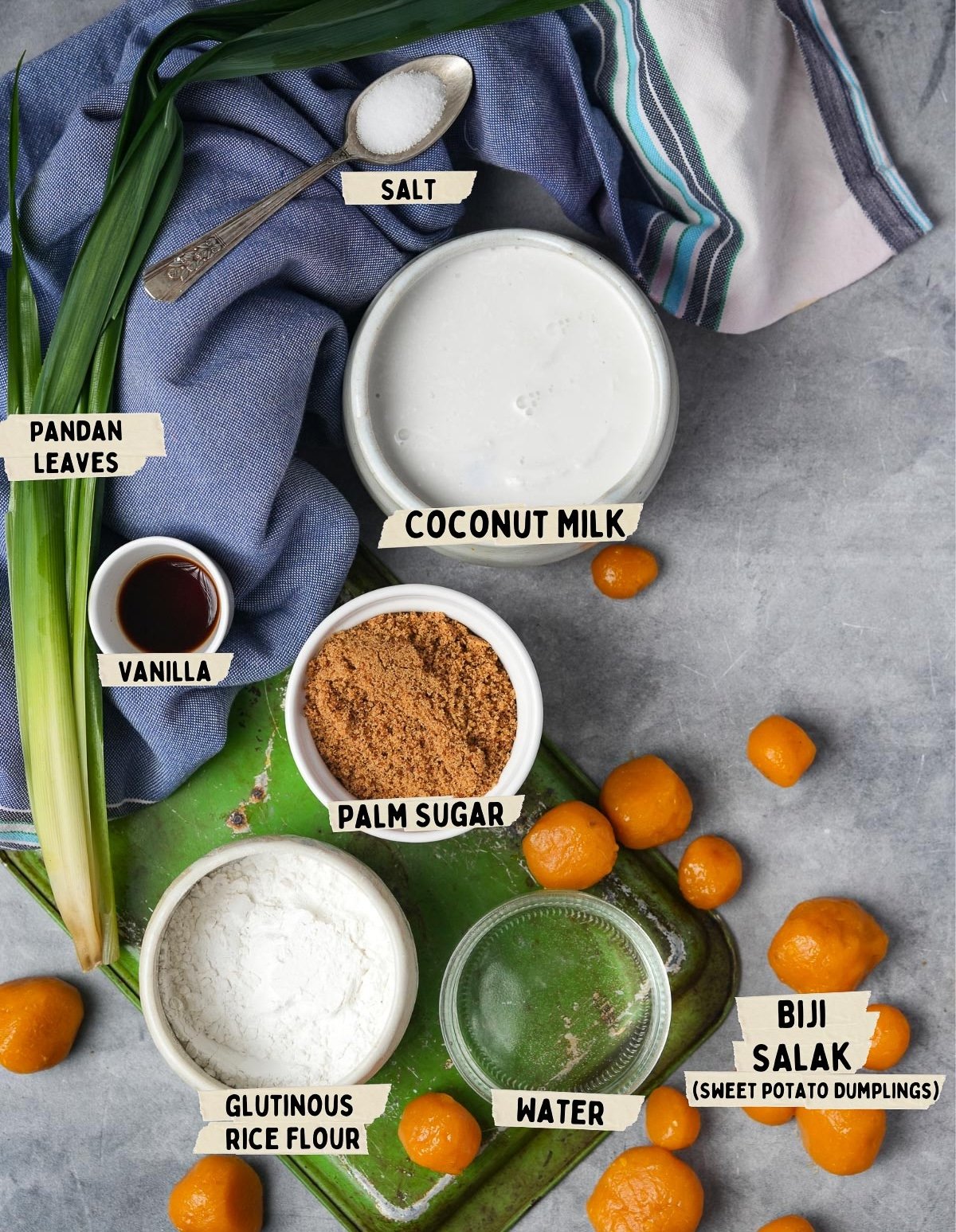 Top view of ingredients for Biji Salak and Bubur Sumsum, including pandan leaves, salt, coconut milk, vanilla, palm sugar, glutinous rice flour, water, and sweet potato dumplings arranged on a green board.