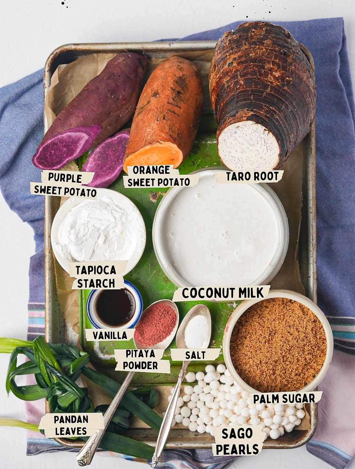 Ingredients for a Bubur Cha Cha dessert are arranged on a tray: purple and orange sweet potatoes, taro root, coconut milk, tapioca starch, vanilla, pitaya powder, salt, palm sugar, sago pearls, and pandan leaves.