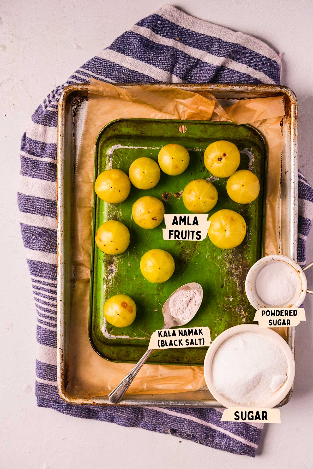 An overhead view of vibrant amla fruits on a green tray accompanied by small, labeled bowls of kala namak, powdered sugar, and sugar. The tray sits elegantly on a striped cloth backdrop.