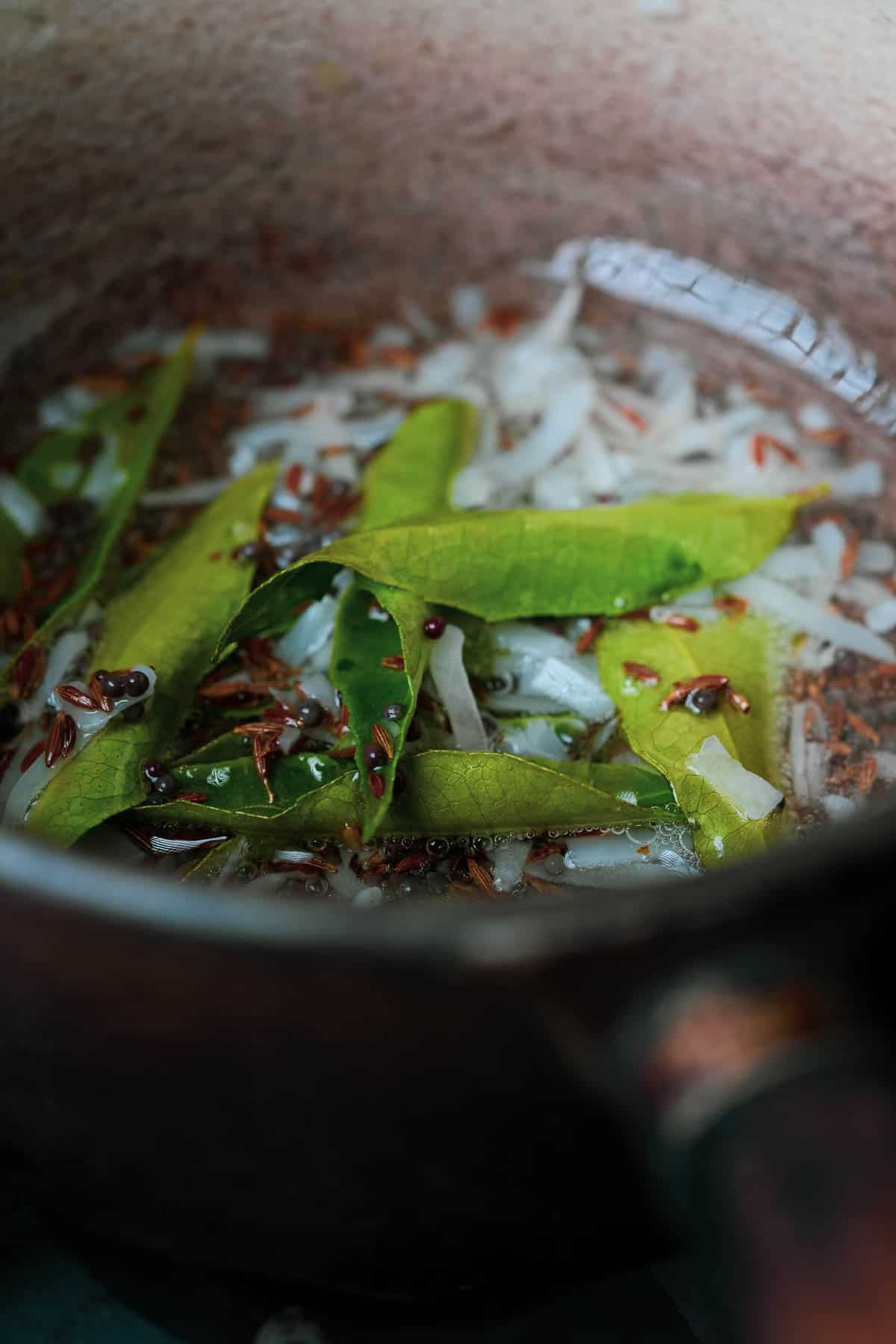 A pan with green curry leaves, coconut, mustard seeds and cumin seeds frrying in it.