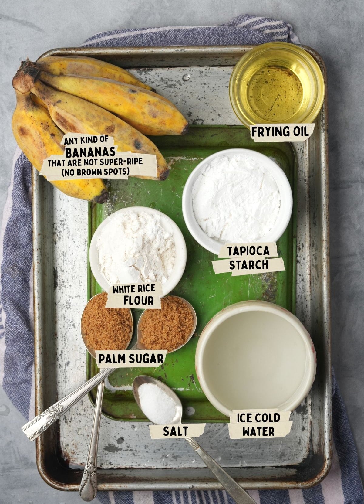 Ingredients for a delicious pisang goreng recipe are laid out, labeled as: bananas, frying oil, tapioca starch, white rice flour, palm sugar, salt, and ice-cold water.