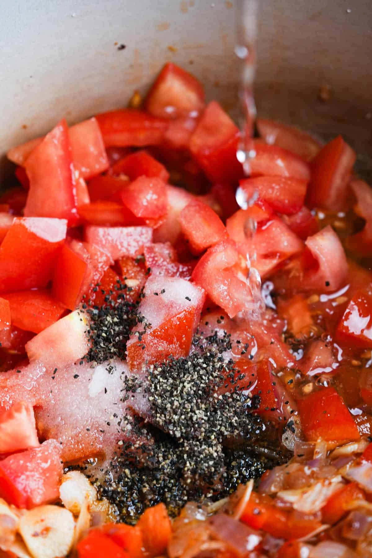 A pot with tomatoes, peppers, and spices has water being poured into it.