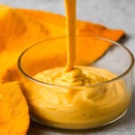 A thick, creamy yellow plant-based cheese sauce being poured into a transparent glass bowl, with an orange cloth partially visible beside it.