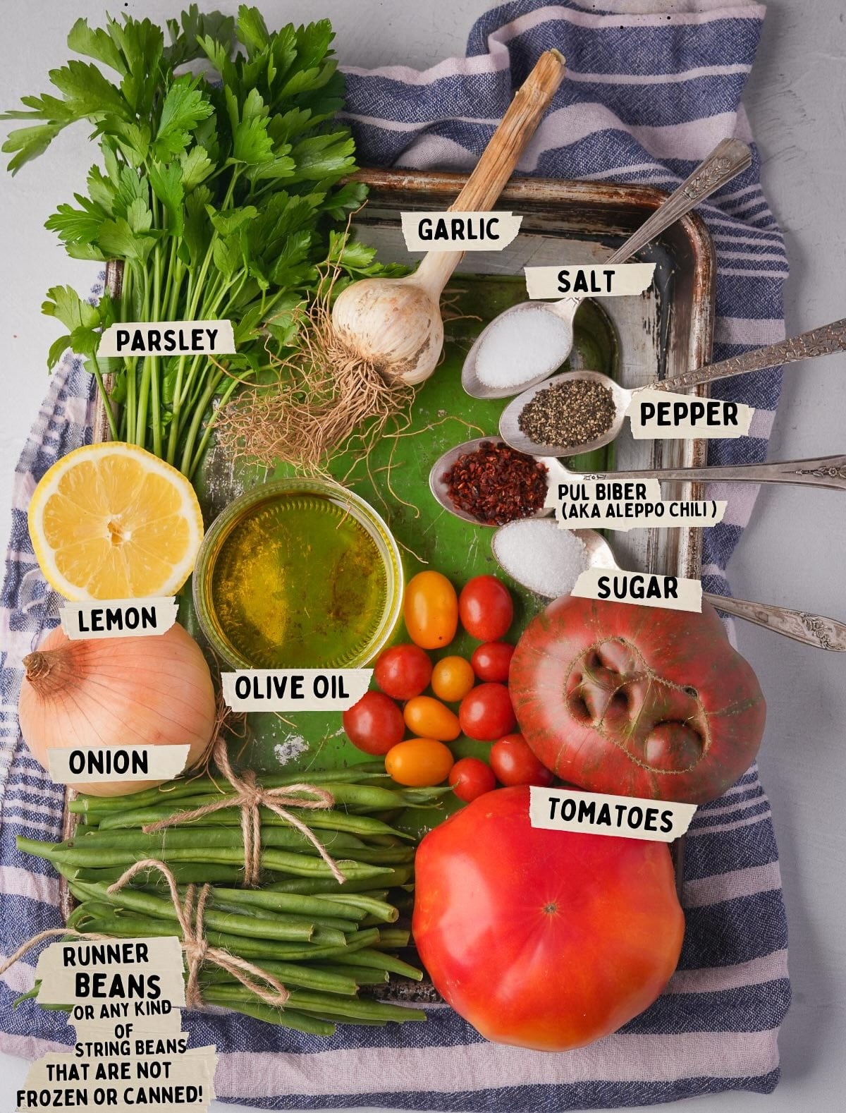 A tray holding various ingredients for making Taze Fasulye Tarifi : parsley, garlic, salt, pepper, pul biber (Aleppo chili), sugar, tomatoes, taze fasulye (runner beans), lemon, onion, olive oil, and cherry tomatoes on a striped cloth.