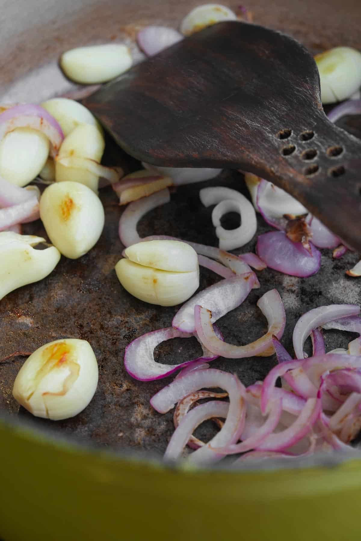 A wooden spoon is being used to stir shallots and garlic in a dry pan.