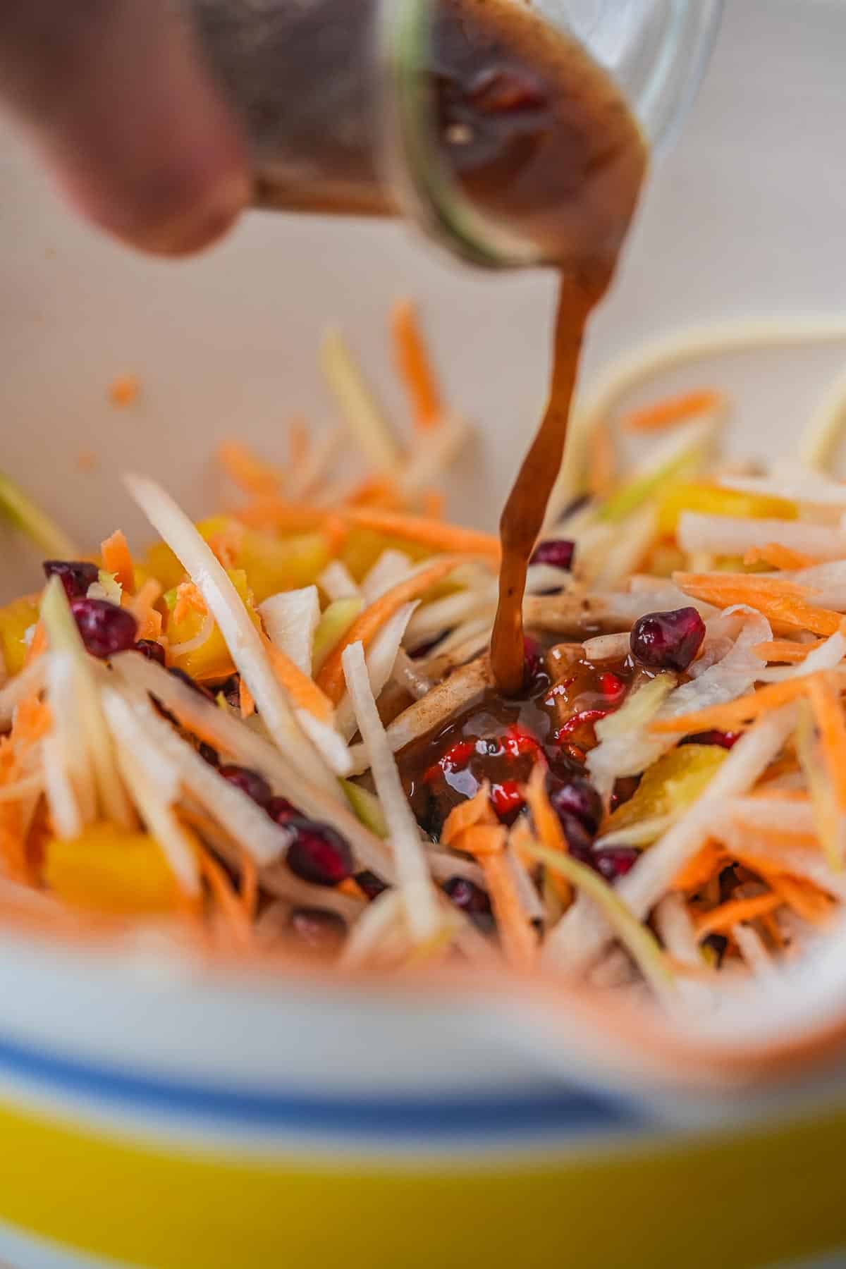 A person is pouring a dressing into a bowl of prepped veggies and fruits.