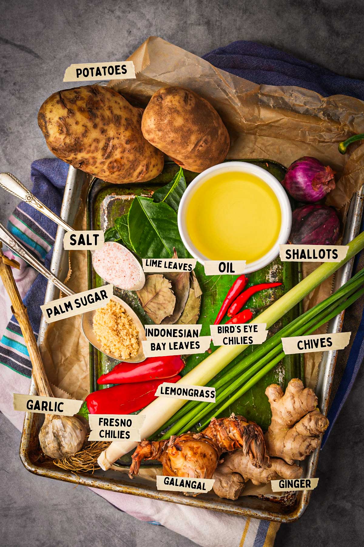 A tray with kentang, shallots, chives, garlic, ginger, galangal, lemongrass, chilies, lime leaves, bay leaves, palm sugar, salt and oil. A striped cloth is partially visible.
