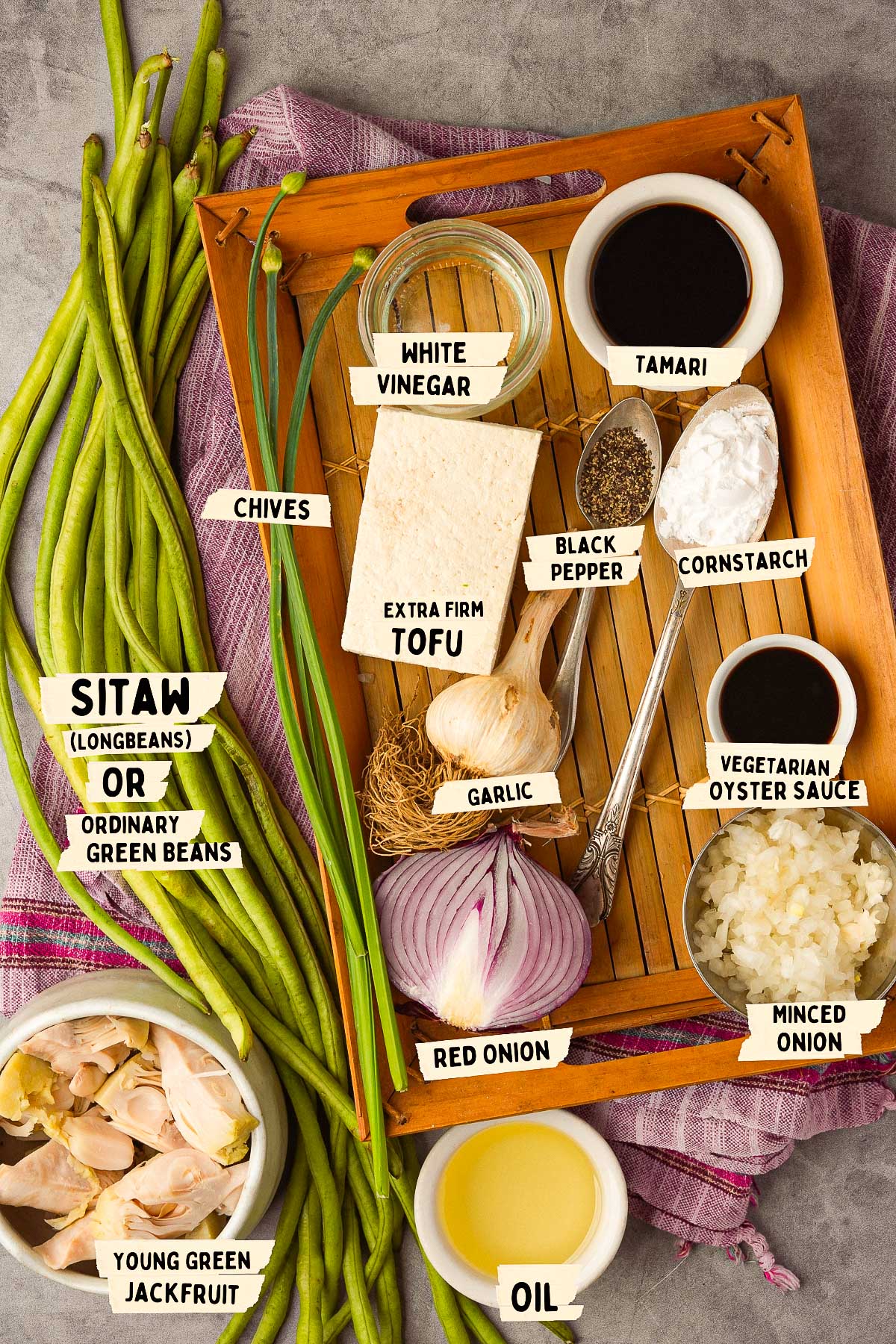 Ingredients for an adobong sitaw recipe are arranged on a tray, featuring sitaw (long beans), tofu, jackfruit, white vinegar, tamari, chives, cornstarch, garlic, red onion, vegetarian oyster sauce, minced onion, black pepper, and oil.