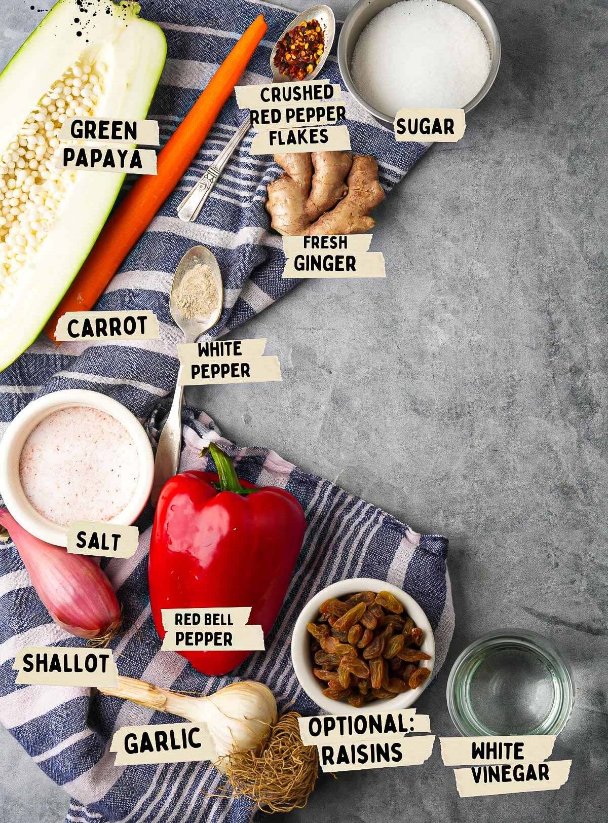Top-down view of various labeled ingredients on a cloth, perfect for making Achara, including green papaya, carrot, shallot, fresh ginger, red bell pepper, optional raisins, garlic, white vinegar, and various spices.