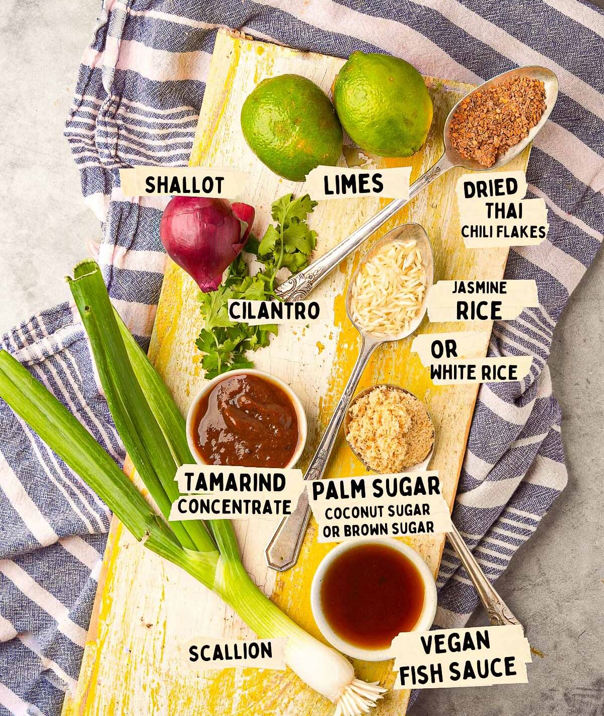 Ingredients for a Thai nam jim jaew recipe include shallot, limes, dried Thai chili flakes, jasmine rice, cilantro, tamarind concentrate, Nam Jim sauce, palm sugar, scallion, and vegan fish sauce arranged on a yellow surface.