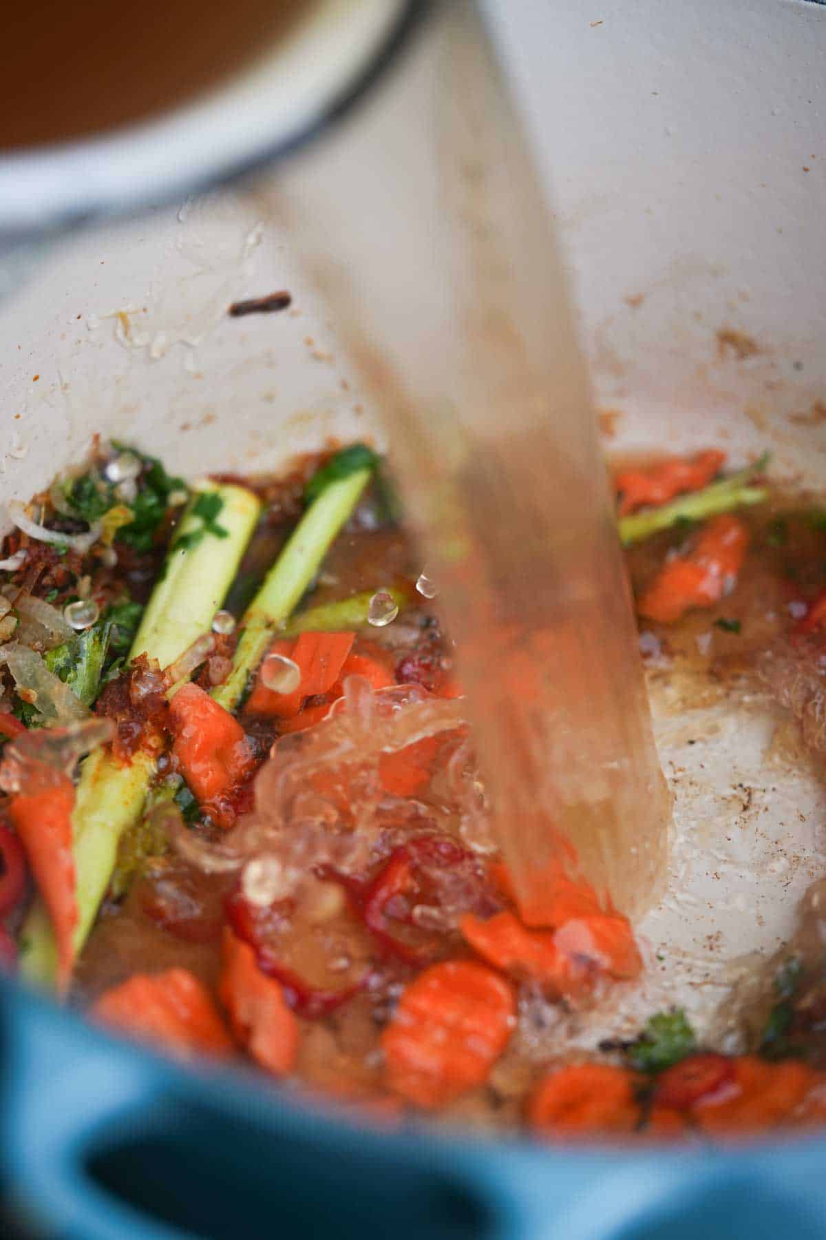 A pot of vegetable broth is poured into a pot of sauteeing herbs and veggies.