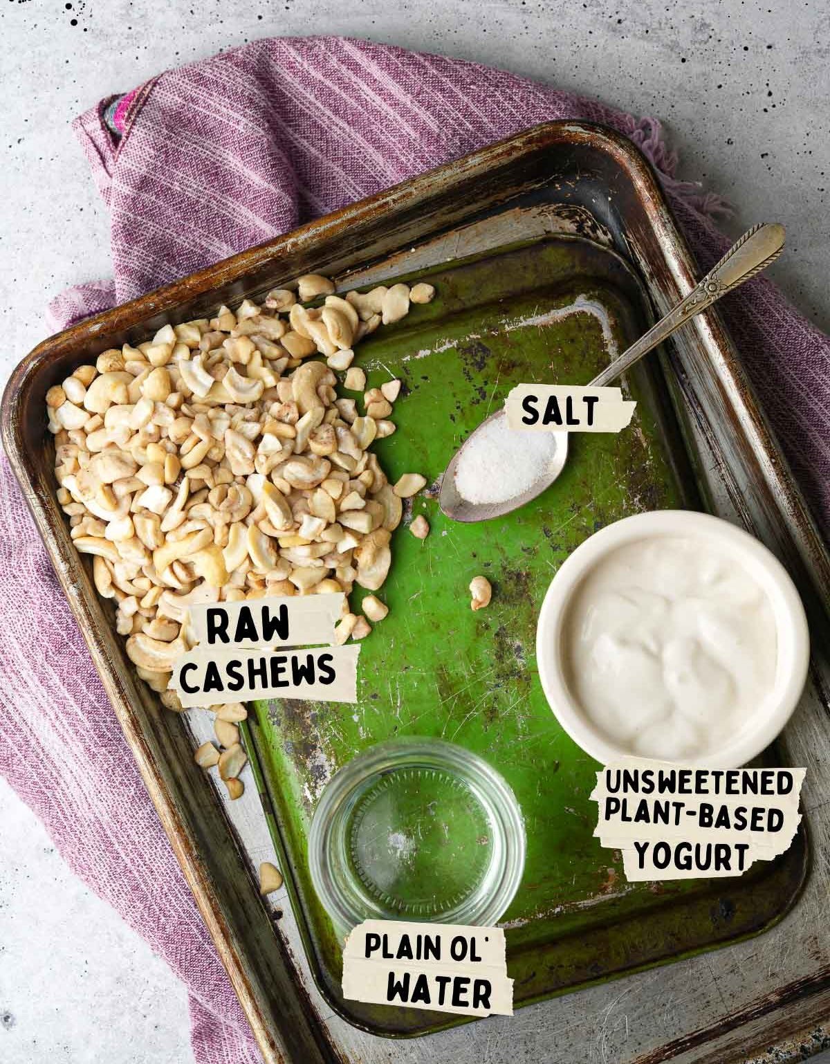 A tray with raw cashews, a small bowl of unsweetened plant-based yogurt, a pinch of salt, and a glass of plain water—perfect ingredients for crafting a smooth vegan cream cheese.