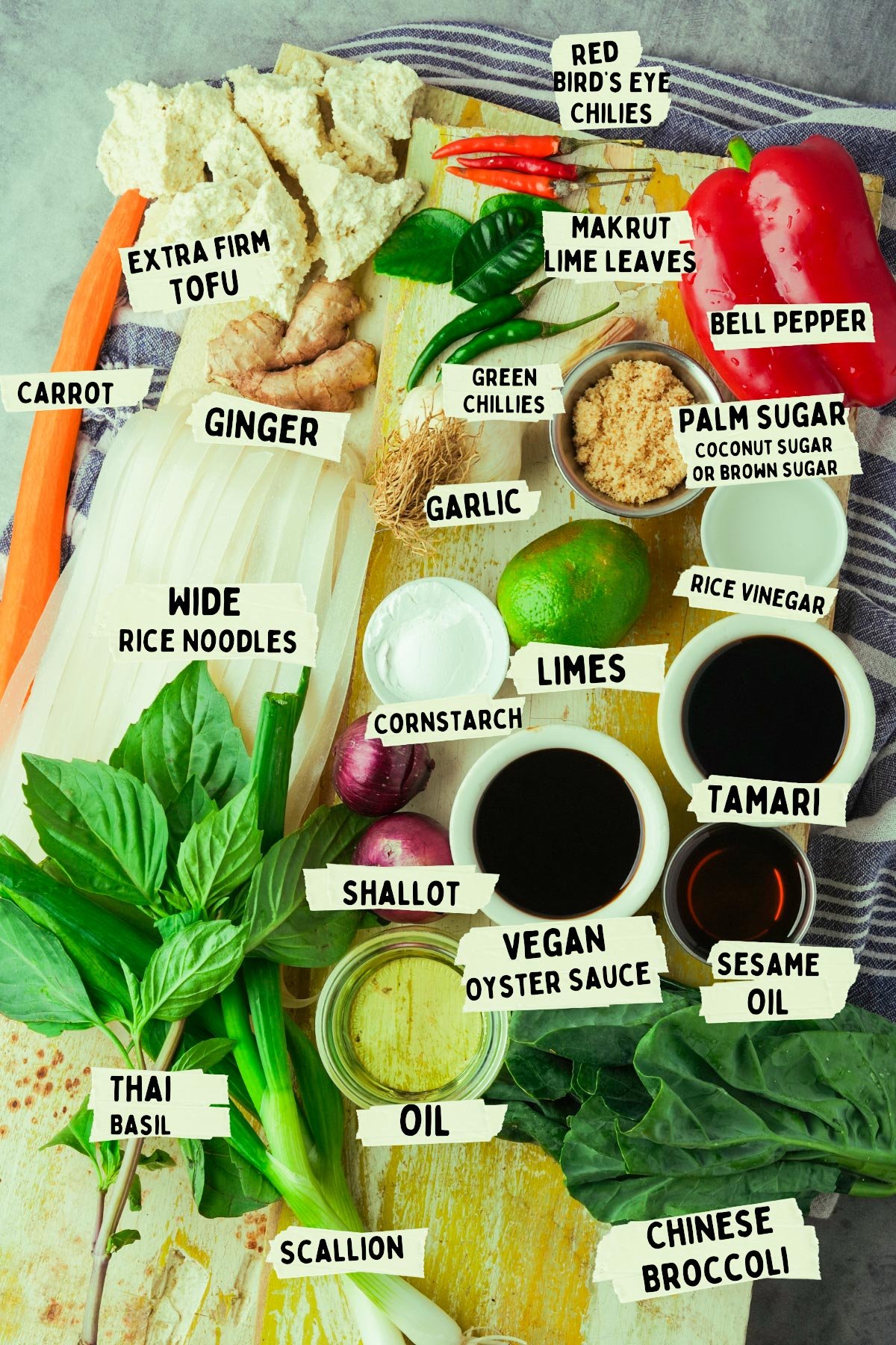 A flat lay of various Asian cooking ingredients, perfect for whipping up Pad Kee Mao, including tofu, rice noodles, vegetables, herbs, and sauces. Each item is beautifully labeled with its name.