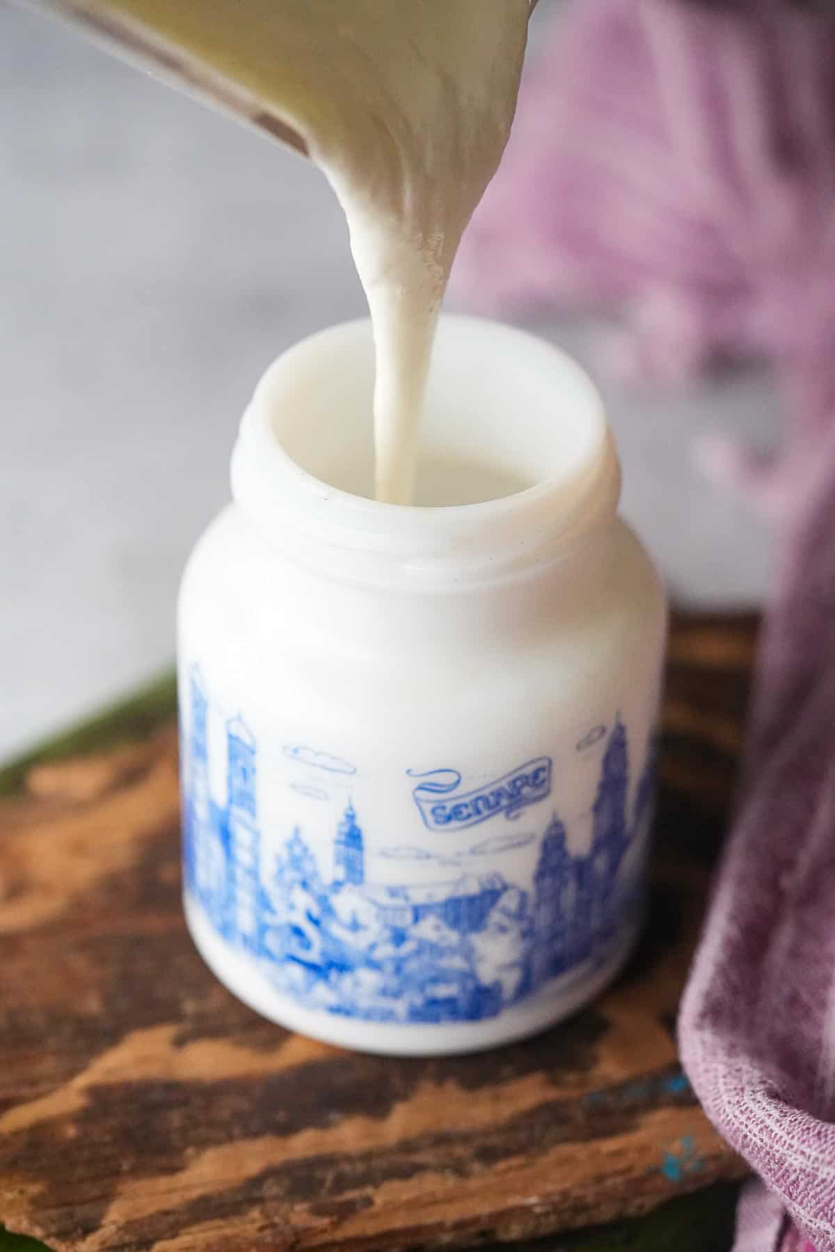 A blender of emulsified vegan sour cream being poured into a jar on a wooden cutting board.