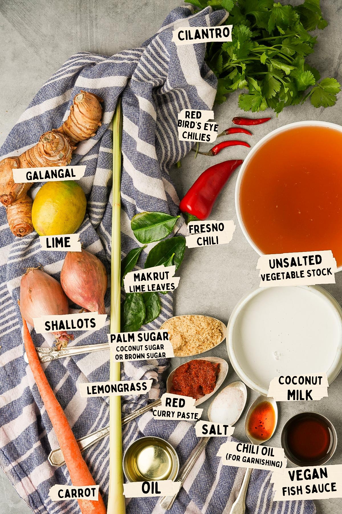 Ingredients for a Tom Kha recipe are artfully arranged on a cloth: cilantro, galangal, lime, shallots, carrot, red chilies, lemongrass, lime leaves, palm sugar, curry paste, salt, oil, chili oil, coconut milk delicately blended with stock and finished with fish sauce.