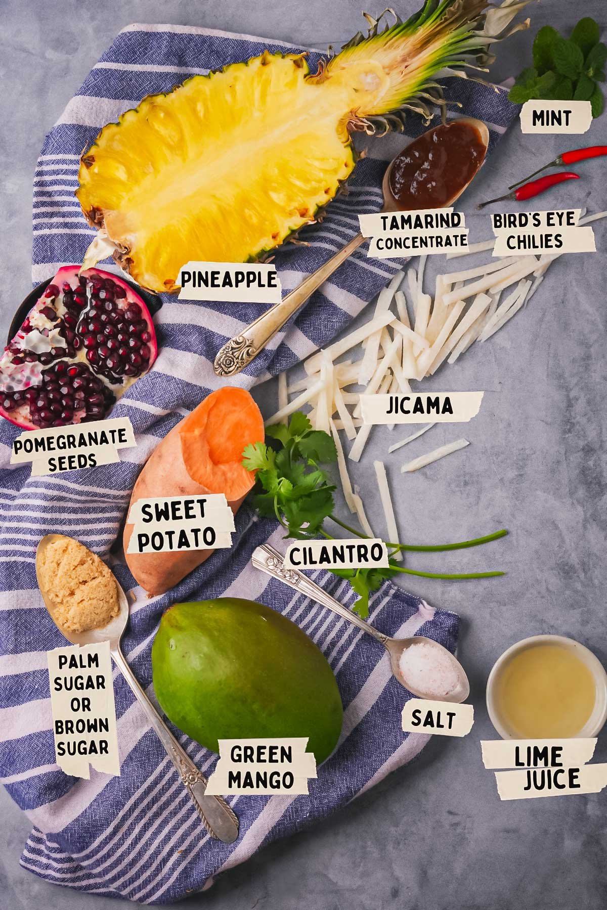 Various ingredients are labeled on a cloth for a delightful Rujak Serut: pineapple, mint, bird's eye chilies, tamarind concentrate, jicama, lime juice, salt, cilantro, green mango, sweet potato, palm sugar, and pomegranate seeds.