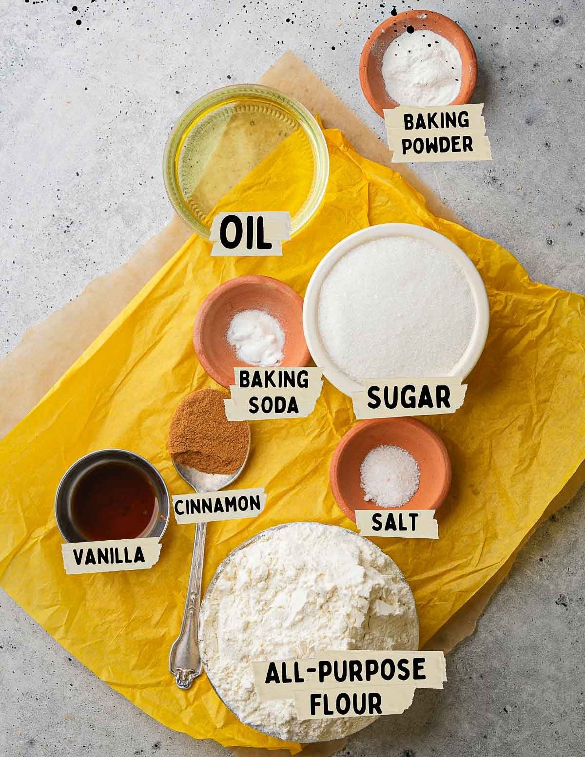 Baking ingredients for buñuelos on a counter include oil, baking powder, sugar, baking soda, salt, vanilla, cinnamon, and all-purpose flour, each neatly labeled in individual bowls and containers.