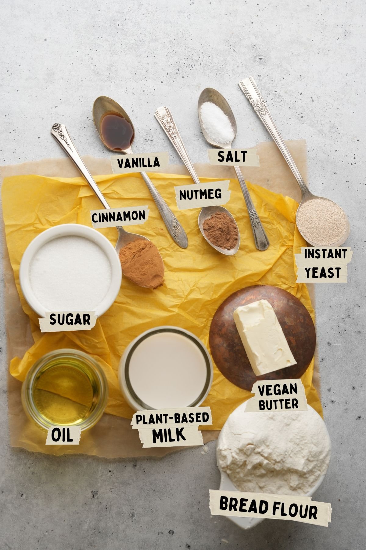 Ingredients for baking a twisted doughnut recipe are labeled on parchment paper and include sugar, vanilla, salt, instant yeast, cinnamon, nutmeg, oil, plant-based milk, vegan butter, and bread flour.