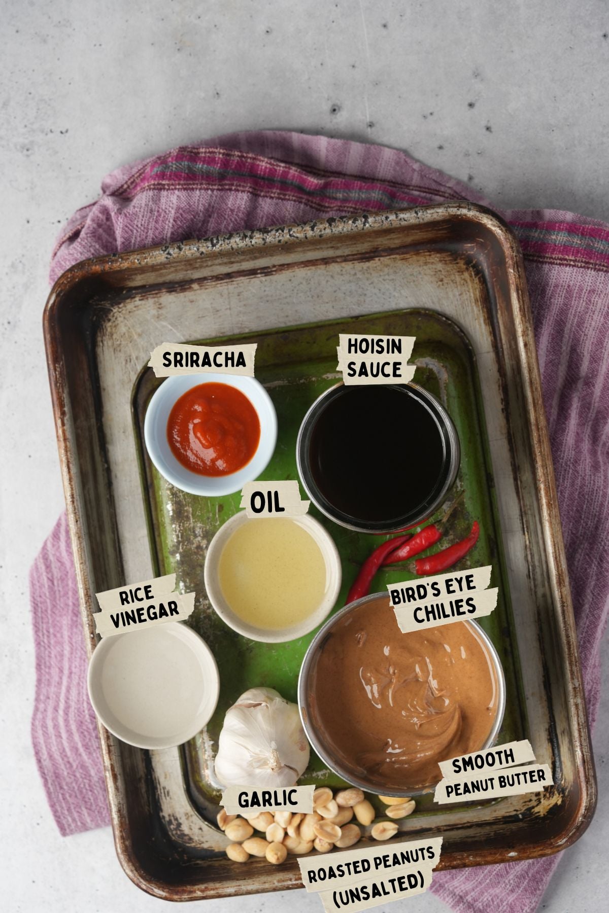 A baking tray with six bowls of ingredients labeled as sriracha, hoisin sauce, oil, rice vinegar, smooth peanut butter, and bird's eye chilies-perfect for making a delectable Vietnamese peanut sauce. Also shown are garlic cloves and roasted peanuts.