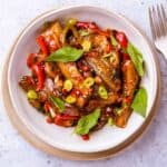 A bowl of stir fried Thai basil eggplant garnished with chili pepper slices, thai basil leaves and scallions in a bowl with a fork.
