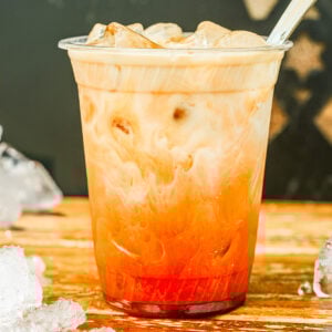 A plastic cup filled with iced Thai tea, showing a gradient of red tea and milk, sits on a wooden surface with ice cubes scattered around—a delightful snapshot of the perfect Thai tea recipe.