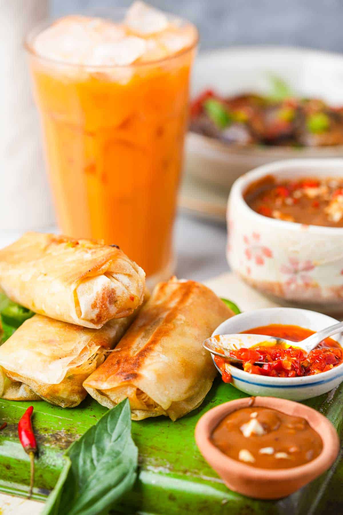 A plate with a spring rolls, peanut sauce, thai tea and thai basil eggplant.
