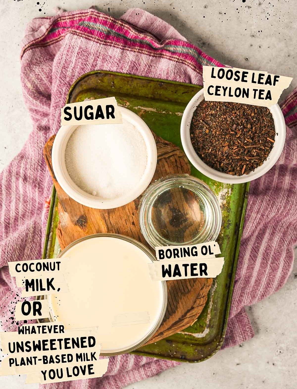 Ingredients for a Thai tea recipe on a tray: bowls of sugar, loose leaf Ceylon tea, water, and plant-based milk, all arranged neatly on a pink cloth.