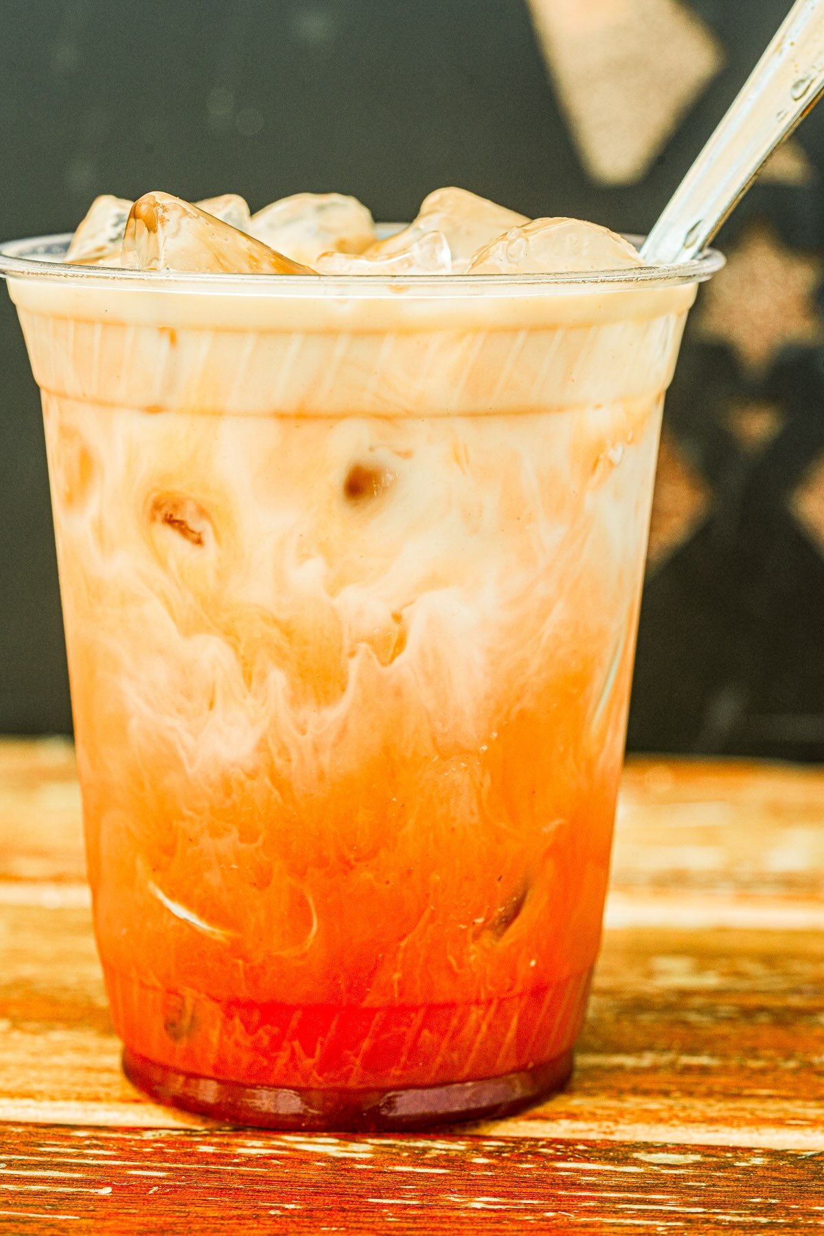 A plastic cup filled with iced Thai tea, showing layers of creamy white and orange colors, sits on a wooden surface with a dark background.