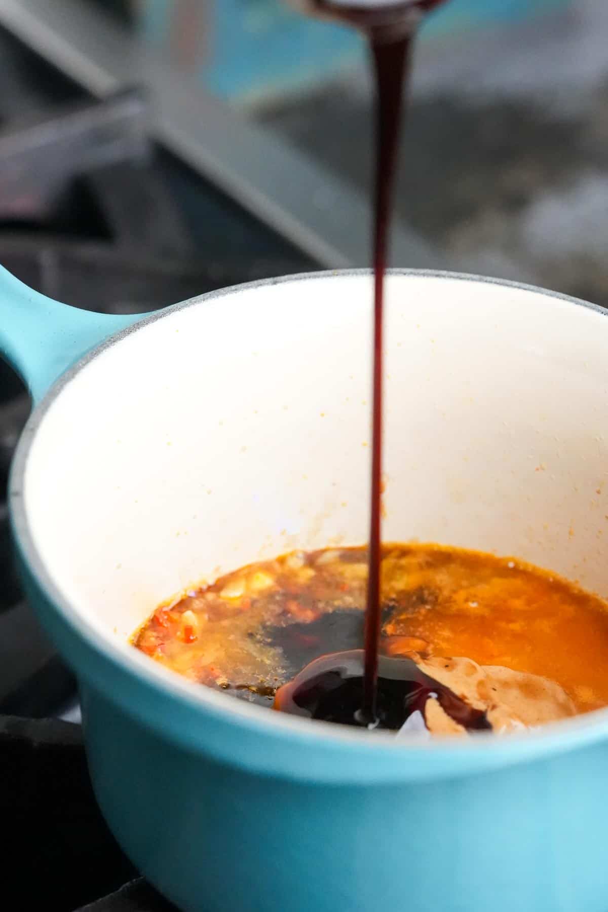A hoisin sauce is being poured into a blue pot containing peanut butter, sautéed garlic and other ingredients.