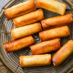A wire rack holds ten golden-brown fried Thai spring rolls.