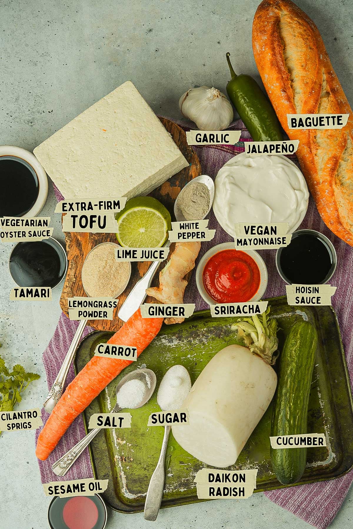 A variety of labeled ingredients on a table, including extra-firm tofu, baguette, carrots, daikon radish, cucumber, vegan mayonnaise, ginger, garlic, jalapeño, lime juice, sauces, and spices-all the essentials for crafting a delicious Vegan Banh Mi.