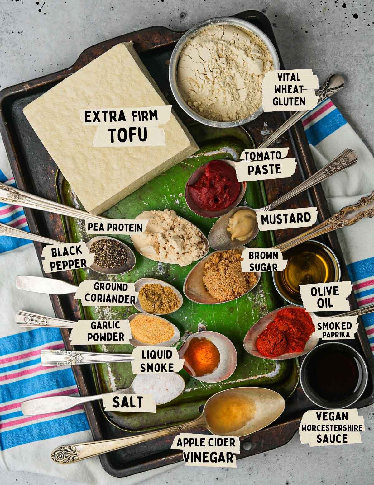 Ingredients for vegan hot dogs are arranged on a tray and labeled. These include extra firm tofu, vital wheat gluten, pea protein, tomato paste, mustard, spices, oil, and other condiments.