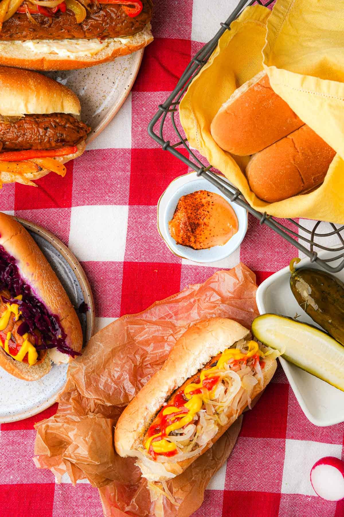 A red and white checkered tablecloth with a spread of vegan hot dogs, condiments and pickles.