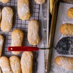 A tray of vegan ladyfingers with powdered sugar and a red spatula.