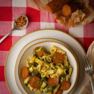 A bowl of pasta with vegan pepperoni and vegetables on a red and white checkered tablecloth.