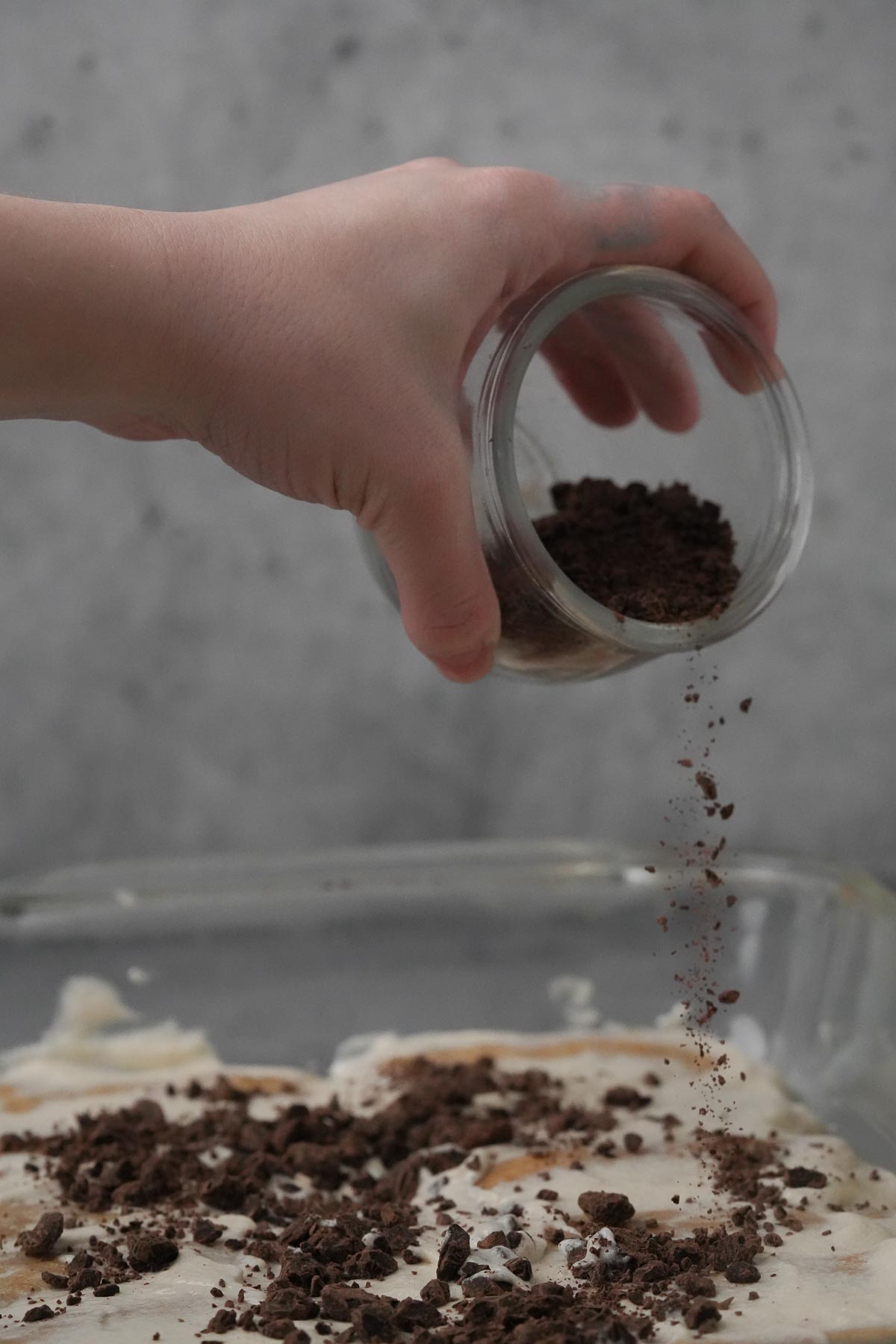 A hand is sprinkling chopped chocolate on a baking dish filled with vegan tiramisu.