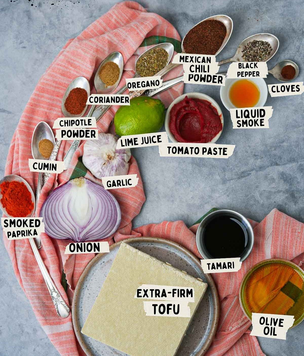 Ingredients for vegan chorizo crumbles, including extra-firm tofu, onion, garlic, lime juice, tomato paste, tamari, olive oil, and various spices, arranged on a pink cloth.