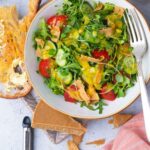 A healthy salad with arugula, tomatoes, cucumbers, Vegan Parmesan Cheese, and a creamy dressing, served with a side of toasted bread.