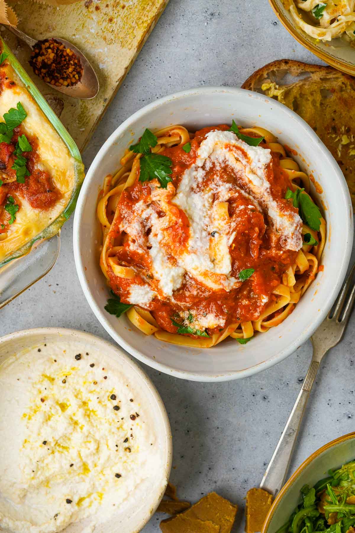 A bowl of pasta with tomato sauce and Vegan Ricotta Cheese, accompanied by various cooking ingredients and utensils.