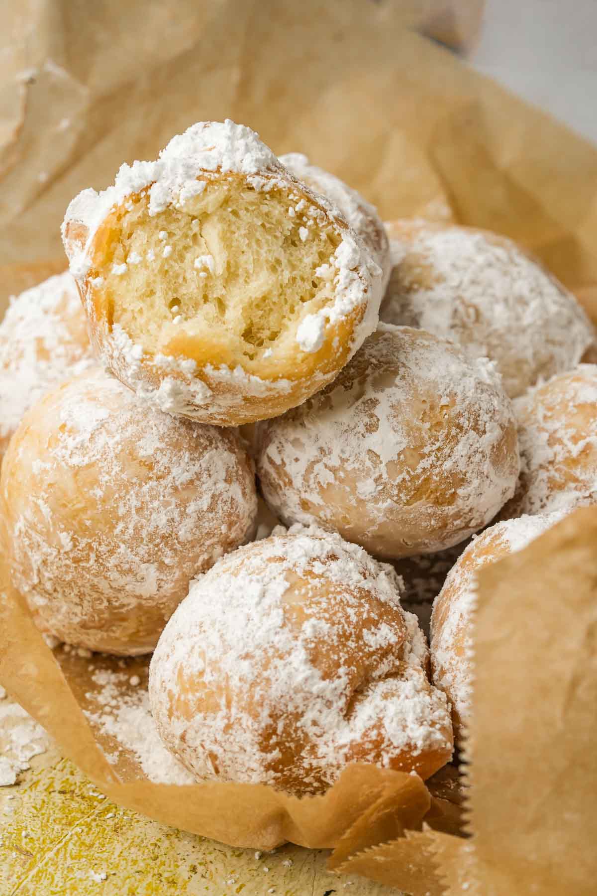 A close-up of powdered sugar-coated Zeppole with a bite taken out of one, revealing a fluffy interior.