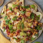 A plate of Indian Aloo Papri Chaat with pomegranate seeds, chickpeas, yogurt, and chutney garnished with herbs.