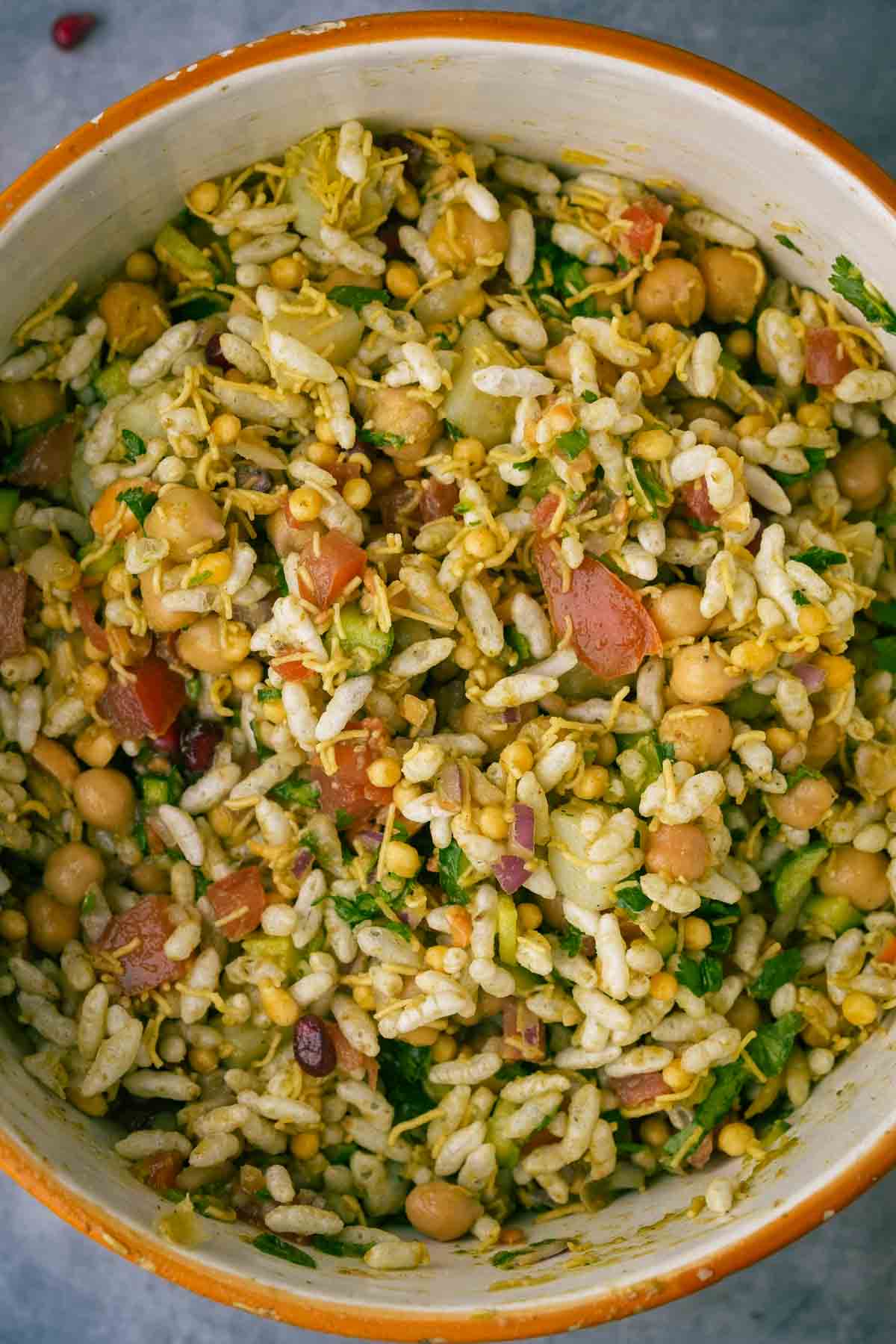 A bowl of mixed bhel puri with chickpeas, tomatoes, cucumbers, and herbs, served in a rustic ceramic bowl.