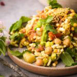 A plate of the vegan Iindian chaat, Bhel Puri, with chickpeas, potatoes, tomatoes, sev, and coriander leaves, served on a textured grey surface.