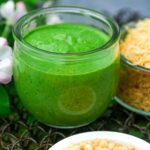 A glass jar filled with fresh green Cilantro Chutney, surrounded by chickpeas and noodles on a textured surface.