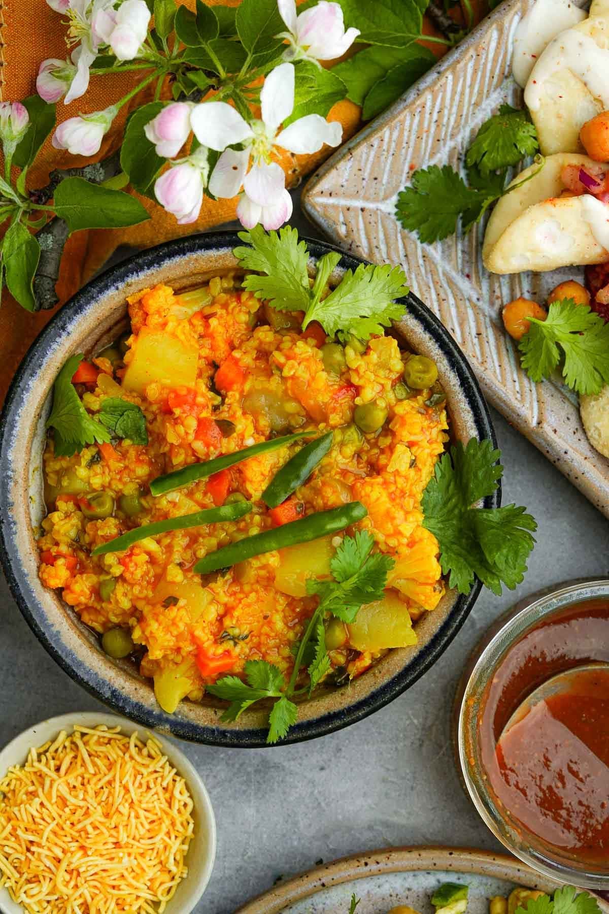 A vibrant dish of daliya curry with vegetables, garnished with fresh herbs, served alongside rice and sauce, with floral accents on a textured grey surface.