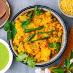 A bowl of daliya khichdi garnished with cilantro and green chilies, served with green chutney and noodle snacks on a gray surface.
