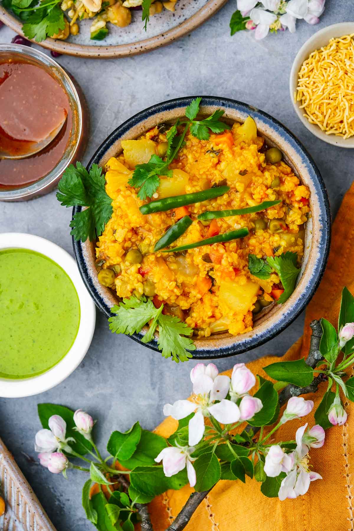 A bowl of indian daliya khichdi garnished with cilantro, served with green chutney, mango pickle, and sev on a gray surface, decorated with white flowers.