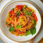 A bowl of spicy hakka noodle stir-fry garnished with sesame seeds, sliced red peppers, green onions, and cilantro.